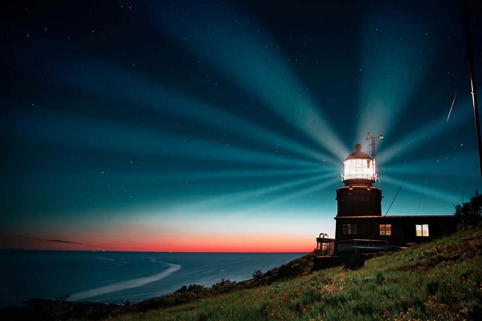 A lighthouse off the coast shining through the night with a sunset off to the far distance
